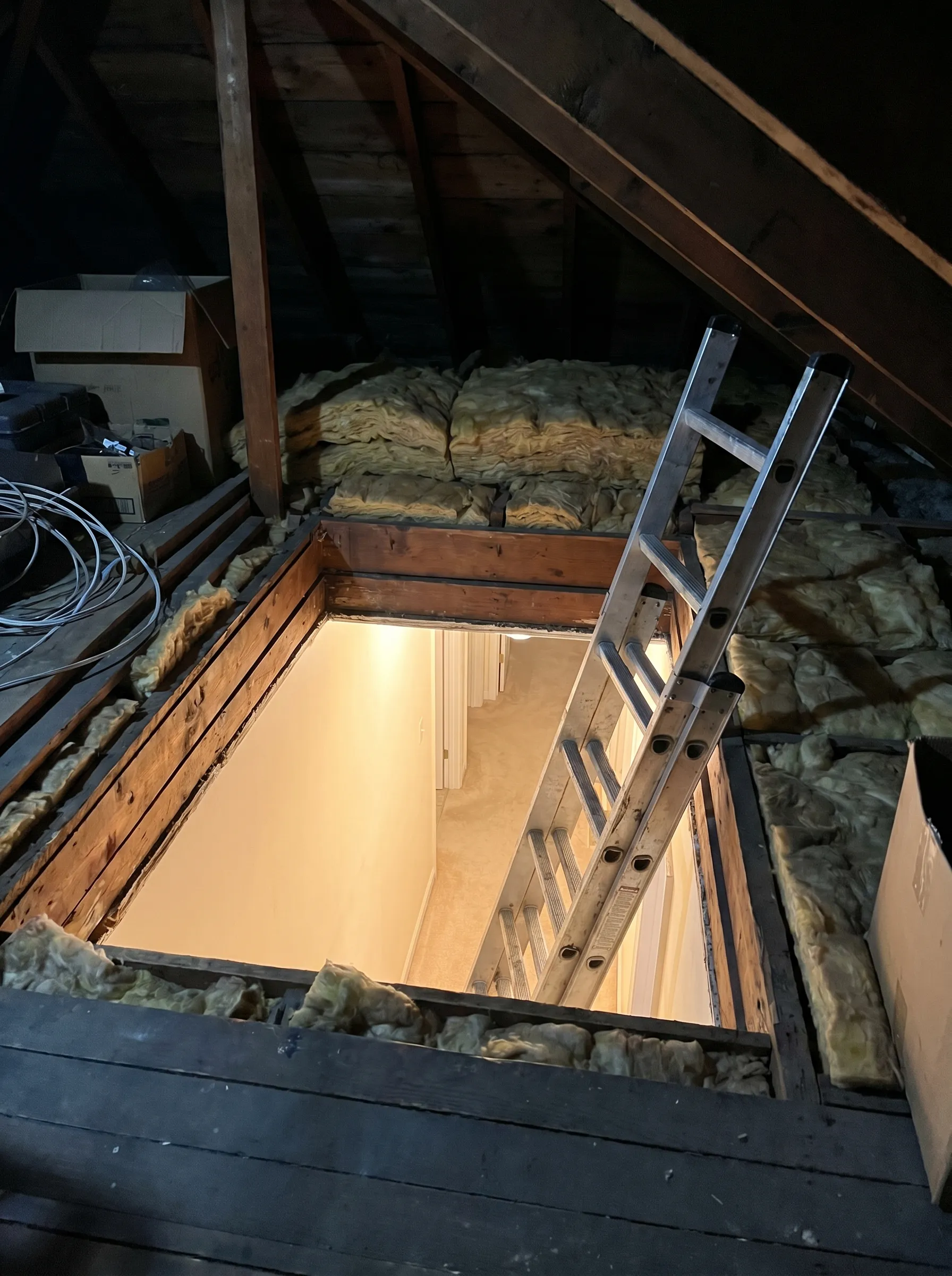 Looking down through an open attic hatch into a warm hallway, ladder in place